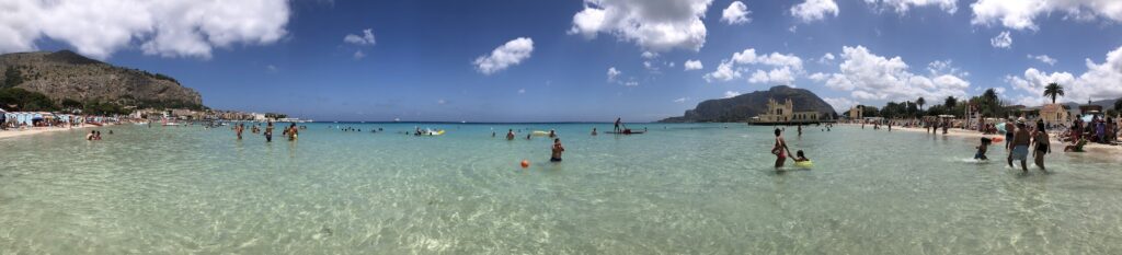 Panoramic view of Mondello beach with greenish blue, crystal-clear water, people swimming, mountains and palm trees in the background and fluffy clouds in a blue summer sky.