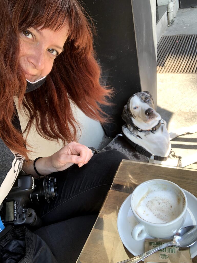 Carol with her Canon camera at a table outside with a cappuccino and frothy milk with a spoon on the saucer. Mucca the catahoula dog sits next to the table waiting for a treat. 