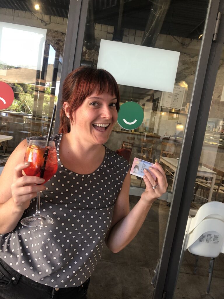 Carol with a big smile in front of a door with a green smiley face sticker on it, holding a Campari spritz in one hand and her permesso di soggiorno (visa) in the other. Finally! 